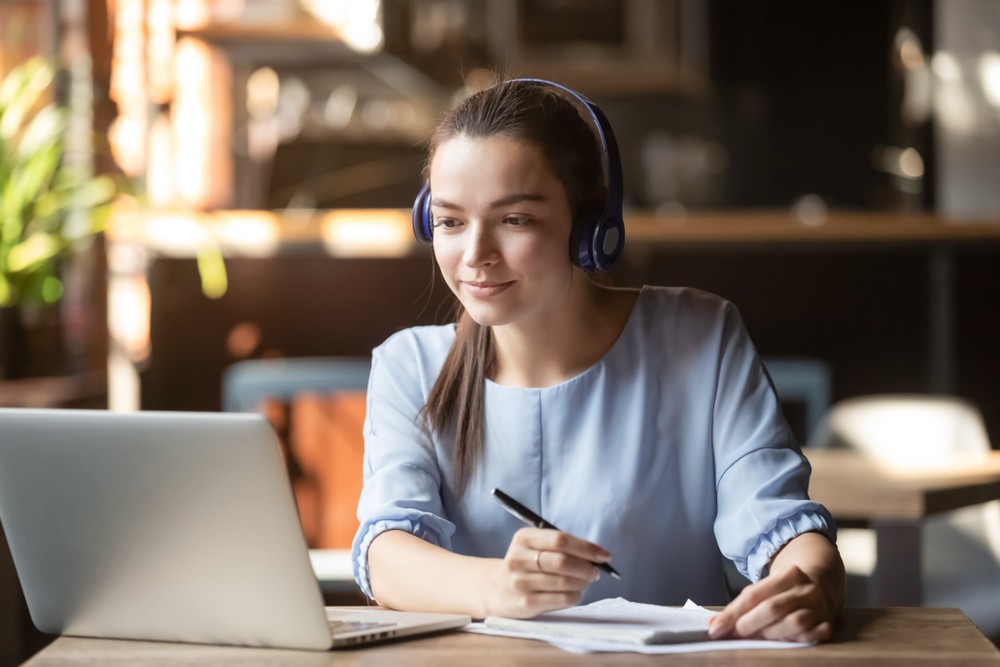 Focused,woman,wearing,headphones,using,laptop,in,cafe,,writing,notes,